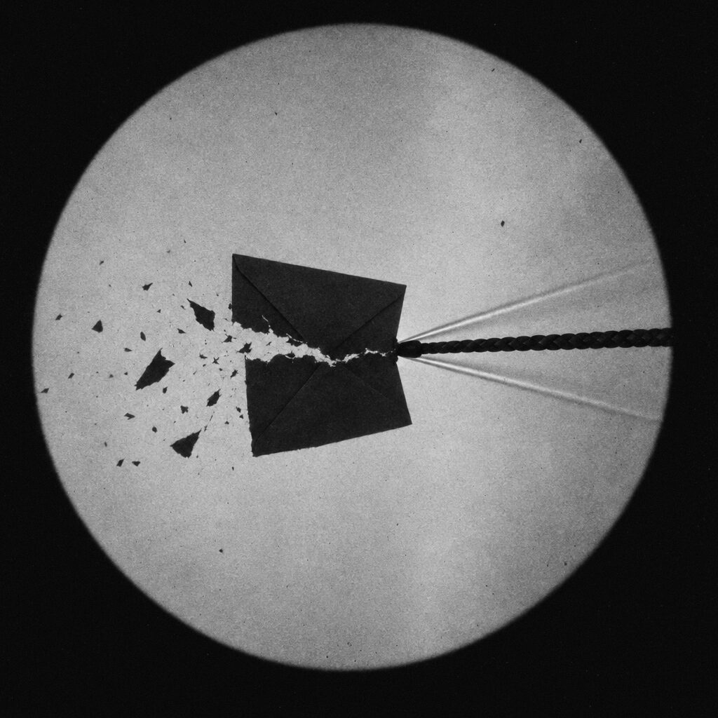 Shadowgraph of a whip tip tearing through a paper envelope mid-air, scraps flying off and a shock cone trailing from the tip.
